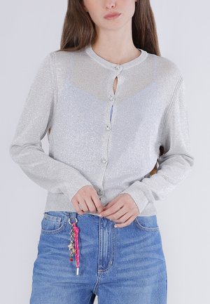 Kvinde iført en sølvfarvet cardigan med knapper over en lys top, kombineret med blå jeans, med en lyserød nøglering fastgjort til jeans.