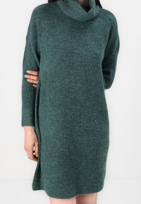 Robe-pull verte en maille avec un col entonnoir haut, des manches longues et des fentes sur les côtés. Texture douce avec un motif côtelé.