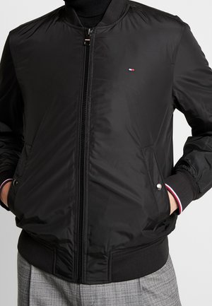 Černá bomber bunda s hladkým, lesklým povrchem, předním zipem, dvěma bočními kapsami a žebrovanými manžetami s červenými a bílými akcenty. Malé logo na hrudi.