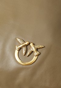 Superfície de pele verde-oliva com um anel em tom de ouro apresentando duas aves detalhadamente elaboradas, com a marca "PINKO" na parte inferior.