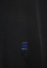 Svart tyg med en slät yta som har en liten blå logotyp i form av en stiliserad "G" och ordet "RAW" nedanför.