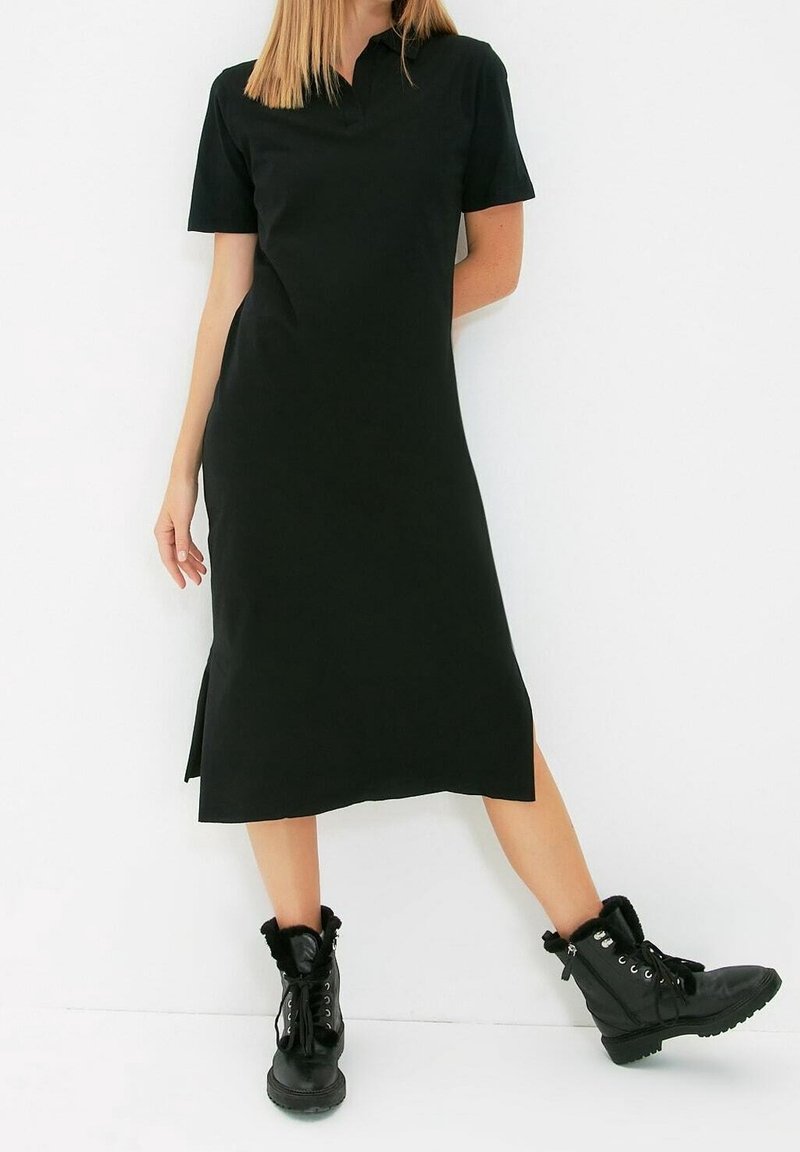 Femme portant une robe midi noire à manches courtes et des bottines noires lacées, debout devant un fond blanc.