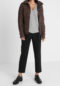 Cardigan zip-up, marrone e nero, top grigio a V, pantaloni neri a sigaretta e sneakers nere con suola spessa.