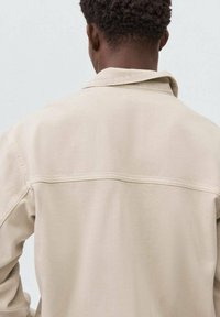 Giacca in tela beige con dettaglio della cucitura a yoke sul retro, colletto appuntito e maniche lunghe. Il tessuto ha una superficie liscia e cuciture visibili.