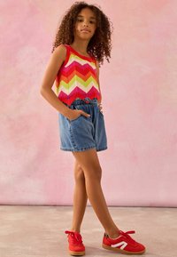 Multikleurige chevron gebreide tanktop met rode accenten; denim shorts met elastische tailleband en zakken; rode sneakers met witte details.