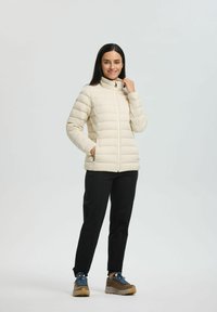 Doudoune beige clair avec des coutures verticales, un col montant et une fermeture éclair sur le devant ; associée à un pantalon noir et des chaussures de randonnée marron.