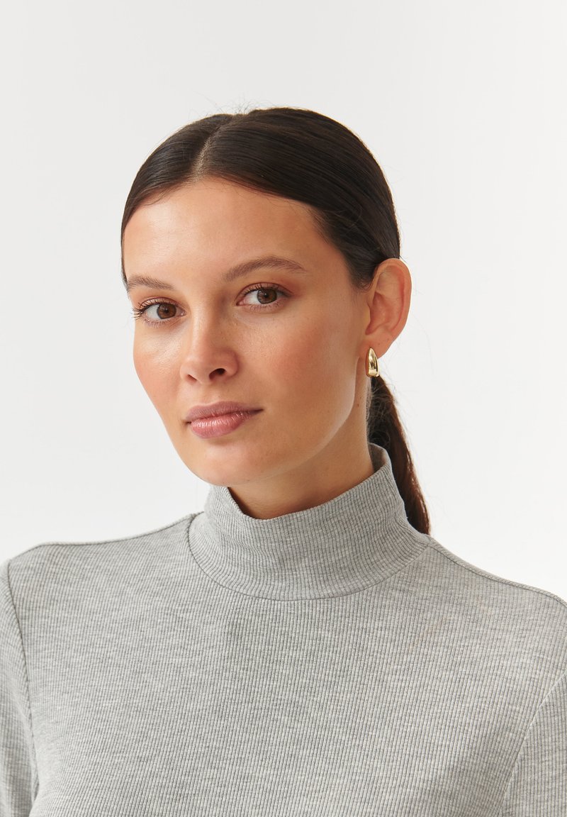 Grauer gerippter Rollkragenpullover mit hohem Kragen, kombiniert mit einem goldfarbenen Creolenohrring. Glatte Textur und minimalistisches Design.
