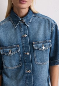 Denim shirt in medium blue with short sleeves. Features two chest pockets with silver buttons and a classic collar. Textured fabric.