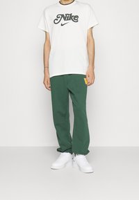 Vit bomull t-shirt med svart Nike-logotyp, tillsammans med gröna sweatpants med en liten gul lapp och vita sneakers.