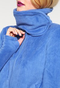 Giacca in pile azzurro chiaro con colletto alto, zip completa e fori per il pollice sulle maniche per una maggiore comodità e calore.