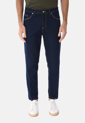 Uomo che indossa jeans aderenti blu scuro, sneakers bianche e camicia verde oliva, in piedi contro uno sfondo chiaro e uniforme.