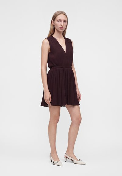 Femme blonde debout portant une robe plissée marron foncé sans manches et des escarpins blancs à petit talon avec bride arrière sur un fond uni gris clair.