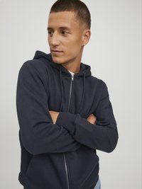 Marineblaues Zip-Hoodie mit Kordelzugkapuze, langen Ärmeln, gerippten Bündchen und glattem Baumwollmix; das Modell posiert mit verschränkten Armen.