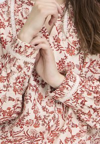 Femme ajustant le poignet d'une blouse crème avec des motifs floraux rouges et de détaillées boutons avec de la dentelle.