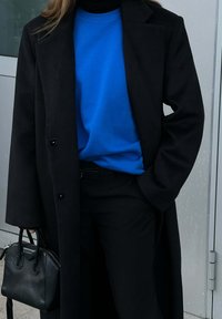 Manteau long noir, sweatshirt bleu vif, pantalon noir et un petit sac à main noir. Détails en bouton et textures lisses visibles.