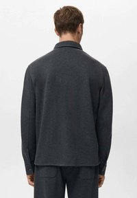 Camicia a maniche lunghe grigio scuro con colletto, caratterizzata da una texture liscia, taglio dritto e polsini con bottoni. Dettagli di cuciture visibili sulle giunture.