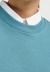 Sudadera de color azul claro con un cuello redondo acanalado, combinada con una camisa blanca de cuello debajo. Textura suave, diseño casual.