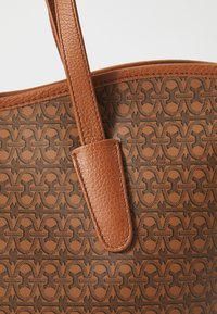 Sac fourre-tout en similicuir marron présentant une surface texturée, un motif géométrique noir et des poignées plates et robustes.