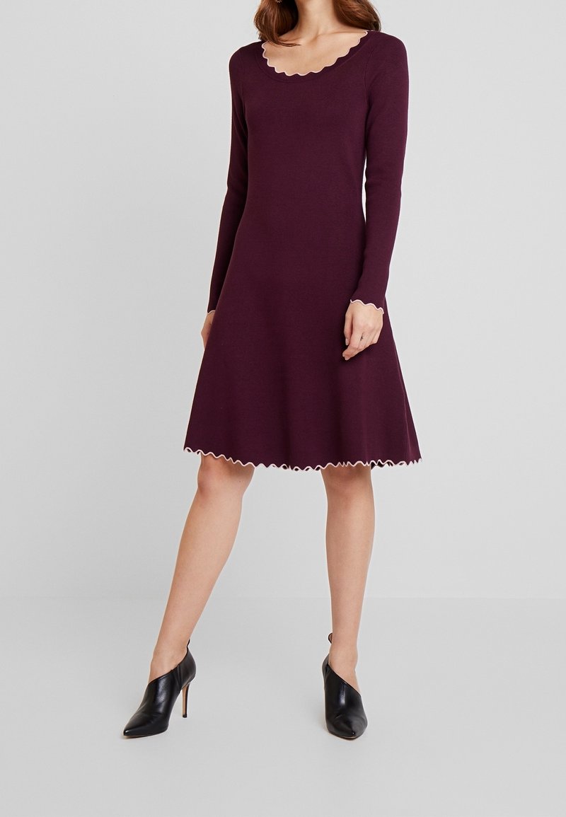Femme portant une robe violette à manches longues, longueur genou, avec des bords festonnés à l'encolure, aux poignets et à l'ourlet, associée à des chaussures à talons noirs.
