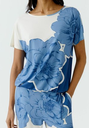 Kobieta ubrana w pasujący niebiesko-biały kwiatowy top z krótkim rękawem i spodnie z dużymi wzorami kwiatowymi, stojąca z jedną ręką w kieszeni.