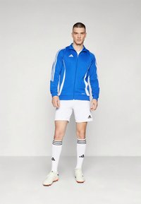 Sinine lukuga sportjakk valgete triipudega, kokku sobitatud valgete lühikeste pükste, mustade triibuliste sokkide ja heledate spordijalatsitega, millel on Adidas'i bränd.