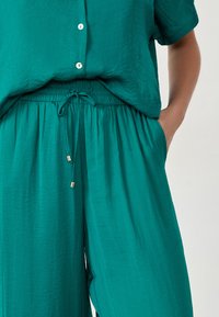 Teal tweed set, bestaande uit een kortekorthalsknopshirt en elastische taillebroek. Het materiaal heeft een zachte textuur en een rijgdetail.