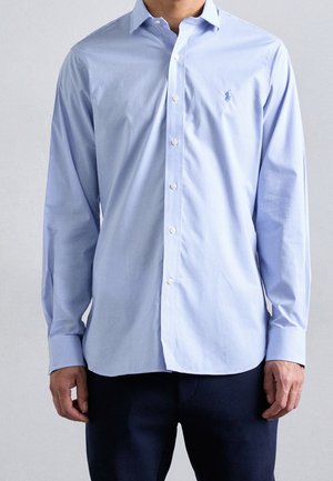 Camisa de dress de cuadros en azul claro, con mangas largas, puños abotonados y un pequeño logotipo bordado en el lado izquierdo del pecho. Textura de tela de algodón.