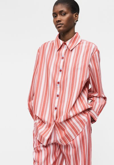 Naine kannab lahtist punase-valge vertikaalastriibulise nööbitavat särki ja sobivaid pükse, seisab käed taskutes tavalise tausta ees.