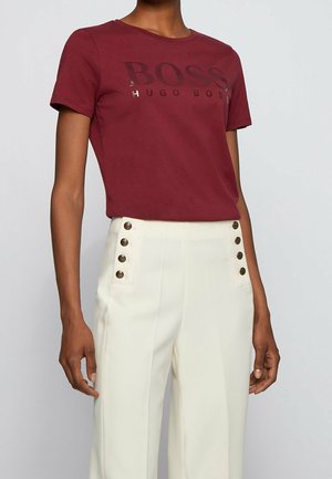 Osoba nosząca bordowy t-shirt Hugo Boss oraz białe spodnie z wysokim stanem, z dwoma pionowymi rzędami po sześć guzików po każdej stronie.
