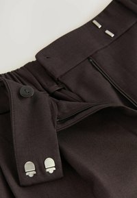 Detailní záběr na pas kalhot tmavě hnědé barvy s knoflíkem, zipem a dvěma stříbrnými kovovými háčkovými zapínáními.