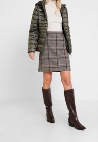 Gewatteerde olijfgroene jas met capuchon, crème coltrui, geruite rok in zwart- en roodtinten, en bruine leren laarzen tot aan de knie met een krokodillenpatroon.