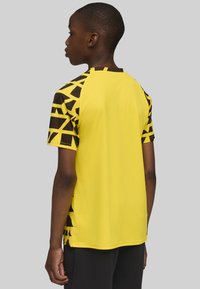 Camisa deportiva amarilla con patrones geométricos negros en las mangas, con una tela texturizada y un escote redondo, cortada para un ajuste relajado.