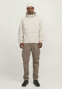 Man i beige huvtröja, taupe cargo-byxor, vit stickad mössa och svarta sneakers, som står mot en enkel ljus bakgrund.