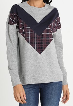 Szara bluza z długim rękawem z granatowo-bordowym wzorem w kratę w kształcie litery V, noszona z niebieskimi dżinsami.