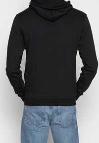 Hombre con una sudadera negra sencilla y vaqueros de mezclilla azul claro visto desde atrás contra un fondo liso.
