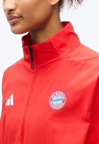 Osoba oblečená v červenej športovej bunde na zips s logom FC Bayern München na hrudi a čiernou sťahovacou zarážkou zipsu.