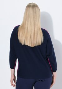 Blonde Person mit glattem Haar, der einen dunkelblauen Pullover mit pinkem Besatz und dunkelblauen Hosen trägt, von hinten vor einem weißen Hintergrund.