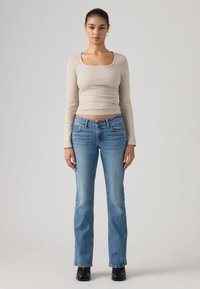 Νεαρή γυναίκα που στέκεται με το πρόσωπο προς τα εμπρός, φοράει μπεζ μακρυμάνικο τοπ, μπλε τζιν, μαύρες μπότες, με τα μαλλιά της δεμένα προς τα πίσω, σε απλό φόντο.