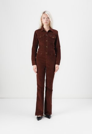 Giubbotto in corduroy marrone con un design aderente, apertura frontale con bottoni, due tasche sul petto e pantaloni a gamba svasata. Indossato con tacchi a spillo neri.