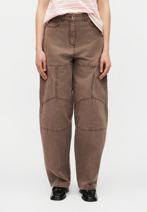 Osoba ubrana w szerokie brązowe spodnie z przeszyciami na kolanach, różową koszulę w paski oraz czarne buty, stojąca na jednolitym tle.