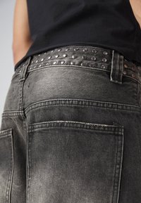 Grå denimjeans har ett nitar prytt svart läderbälte. Det texturerade tyget visar en blekt yta. Bakfickorna är synliga och betonar designen.