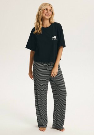 Lächelnde Frau, die ein lockeres schwarzes T-Shirt mit kleinem weißem Hundelogo und schwarz-weiß gestreifte, weite Hosen trägt, barfuß stehend.