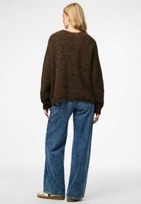 Naine, kes kannab laia pruunist kudumist sviitrit, laia jalgadega siniseid teksaseid ja beeže tennisets, seisab valge tausta vastu, seljaga vaataja poole.