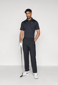 Σετ γκολφ που περιλαμβάνει μαύρο πόλο πουκάμισο, σκουρόχρωμο παντελόνι, λευκά παπούτσια και μαύρο καπέλο. Ο παίκτης γκολφ κρατάει ένα μπαστούνι και φοράει λευκό γάντι γκολφ.