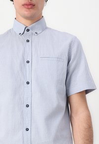 Camicia a maniche corte con bottoni in tessuto azzurro chiaro con un motivo testurizzato, dotata di colletto, taschino sul lato sinistro del petto e bottoni scuri.