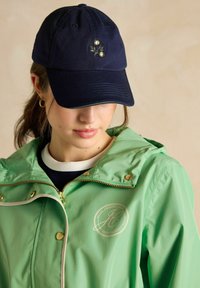 Jūras spēku cepure ar ziedu izšuvumu, apvienota ar piparmētru zaļu ar rāvējslēdzēju no jumta, kas rotāta ar zelta furnitūru un logo detaļām.