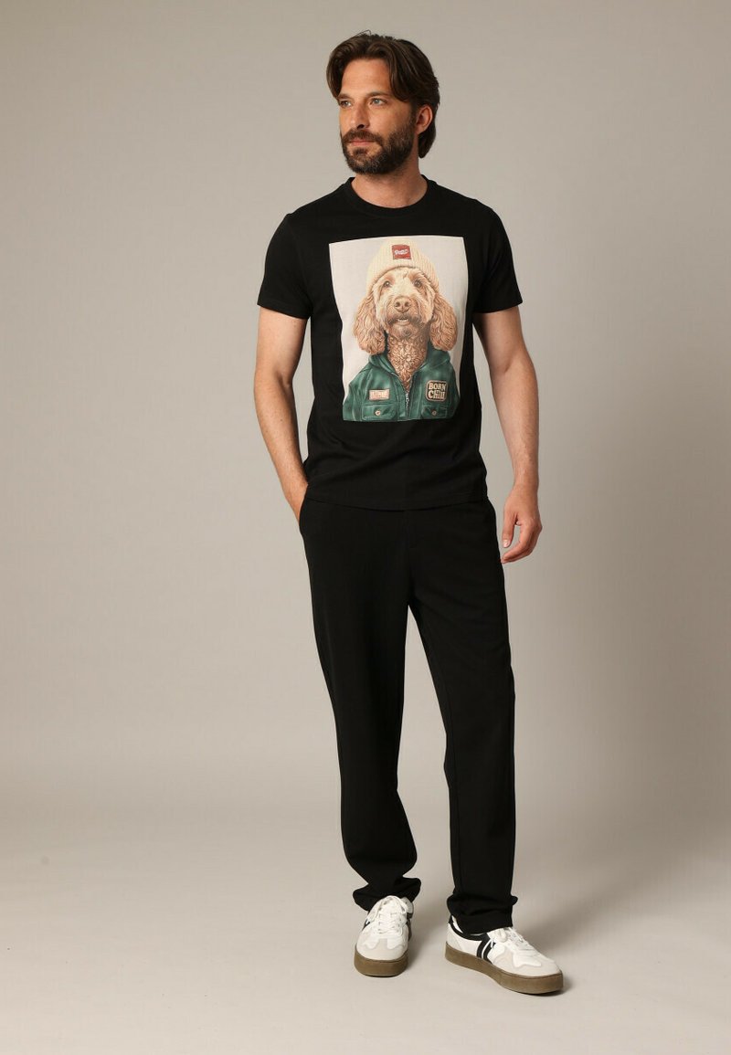 Zwarte T-shirt met een bedrukte grafiek van een hond die een beanie en een jas draagt. Gecombineerd met zwarte broek en witte sneakers.