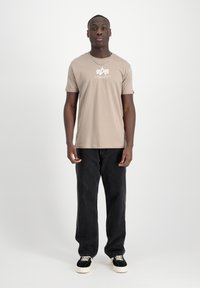 Beigais kokvilnas t-krekls ar baltu logo uz krūtīm, kombinēts ar melniem džinsiem un melnām kedām ar baltām zolēm.