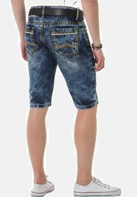 Denim-Shorts in einem ausgewaschenen blauen Farbton mit kontrastierenden gelben Nähten, die bis zur Mitte des Oberschenkels reichen, kombiniert mit einem schwarzen Gürtel und weißen Sneakers.