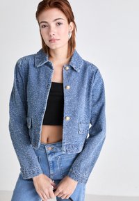 Jeune femme aux cheveux rouges courts portant une veste courte bleue texturée, un crop top noir, un jean bleu taille haute et des boucles d'oreilles en argent.